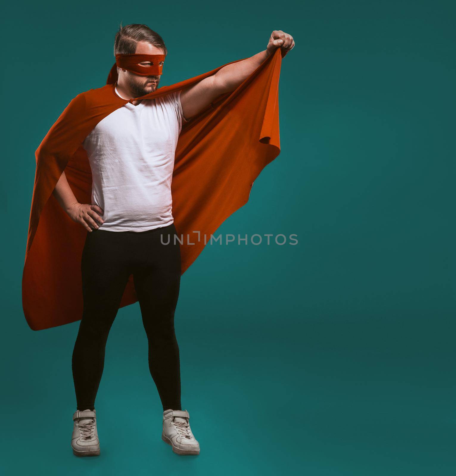 Super hero savior man ready to fly. Legendary brave man in red mask holds his cloak ready to save world from crime. Isolated on biscay green background with text space at right side by LipikStockMedia