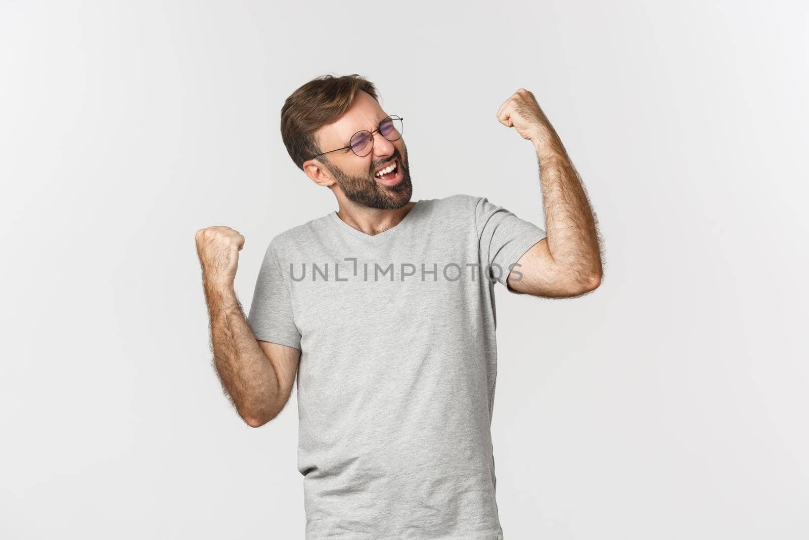 Image of excited man in gray t-shirt and glasses, winning something, rejoicing and feeling like champion, standing over white background.