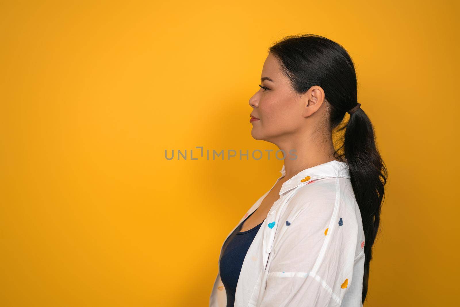 Middle aged woman with perfect skin looks at copy space. Profile view of pretty Asian woman with long hair on yellow background by LipikStockMedia