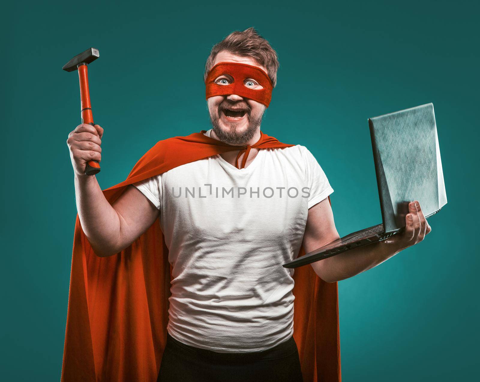 Smiling Super hero man holds hammer and laptop ready to repair the computer. Man In Red Super Hero Uniform posing on biscay green background. Computer repair concept.