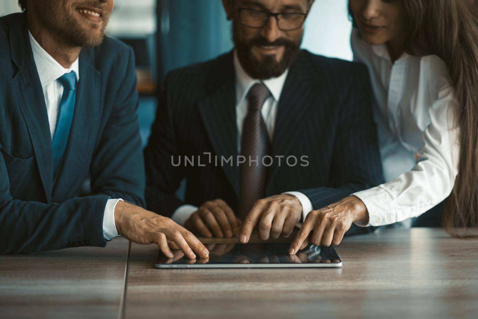 Happy colleagues using the same digital tablet together. Two men and a woman work in office. Cropped image. Selective focus on business people hands. High quality photo.