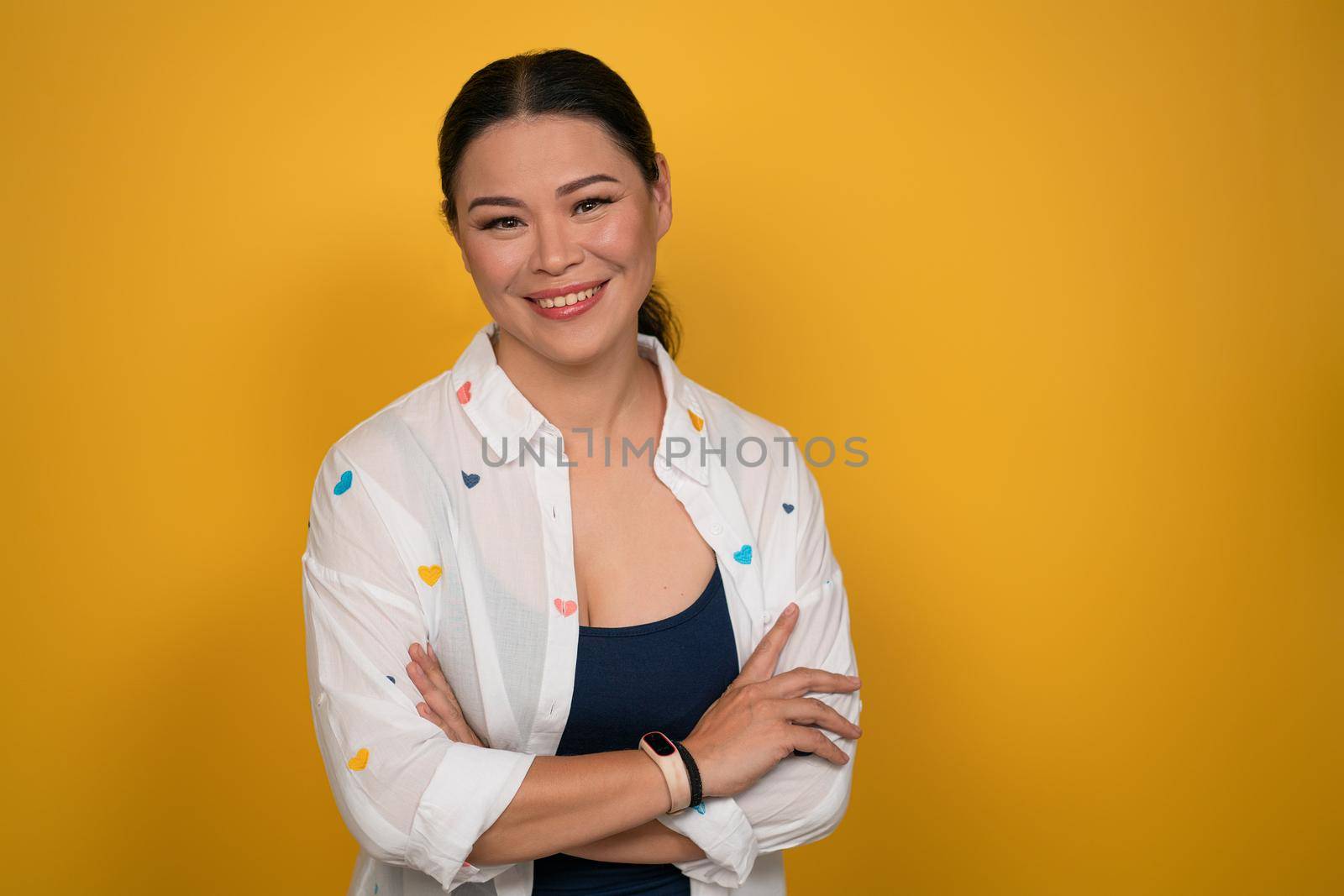 Confident Asian woman smiles standing crossed hands on yellow background. Copy space by LipikStockMedia