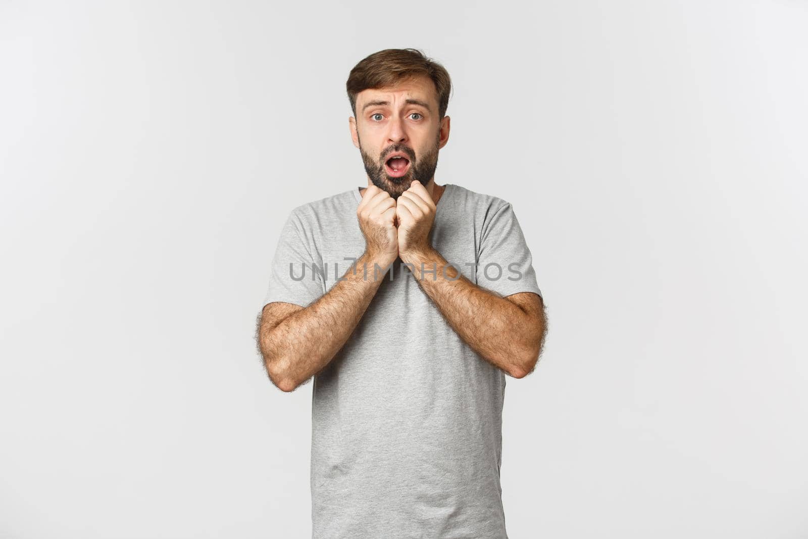 Portrait of compassionate guy with beard, wearing grey t-shirt, gasping and looking worried, standing over white background by Benzoix