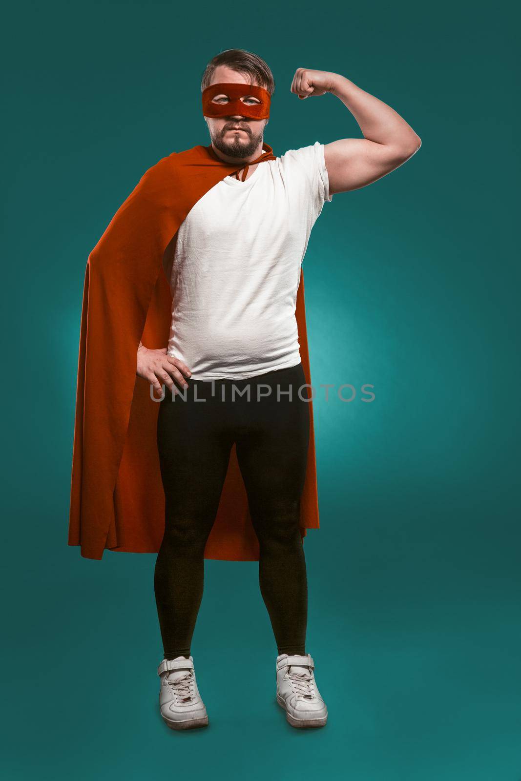 Super Hero Man Shows His Muscles And Ready To Save World. Serious Man In Red Super Hero Mask And Coat Showing His Muscles Looking At Camera. Cut Out On Biscay Green Background.