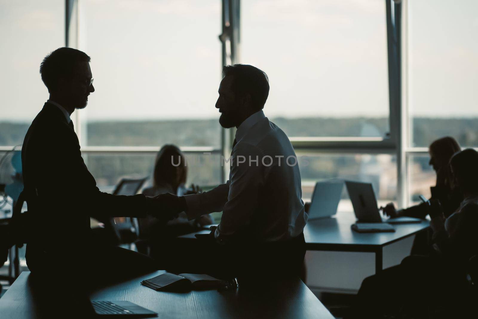 Dark silhouettes of office workers against of window on blurred background. Selective focus on two businessmen communicating while sitting on table in foreground. Toned image.