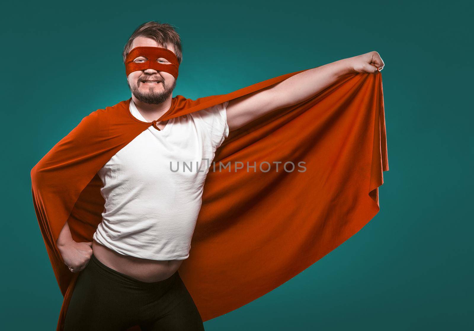 Funny Super Hero Man Ready To Fly Save World. Smiling Man In Mask And Red Superhero Costume Holding His Cloak In His Hand Posing On Biscay Green Background.