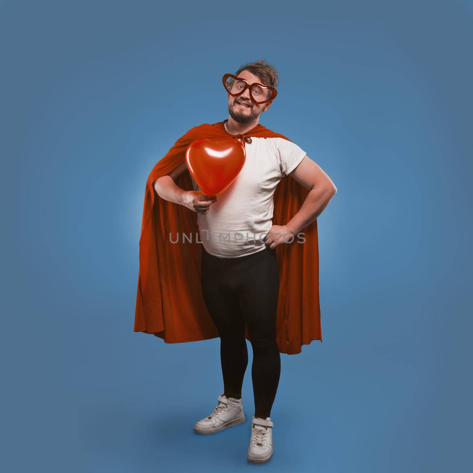 Romantic super hero holds big heart. Happy man in love wearing superhero costume and heart-shaped glasses isolated on blue background. Valentine's day concept. Copy spase on both sides by LipikStockMedia