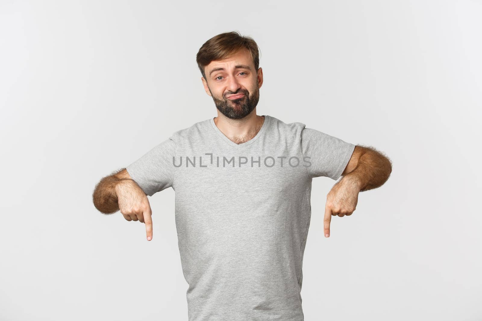 Portrait of unimpressed skeptical guy with beard, wearing gray t-shirt, sulking and pointing fingers down disappointed, standing over white background by Benzoix
