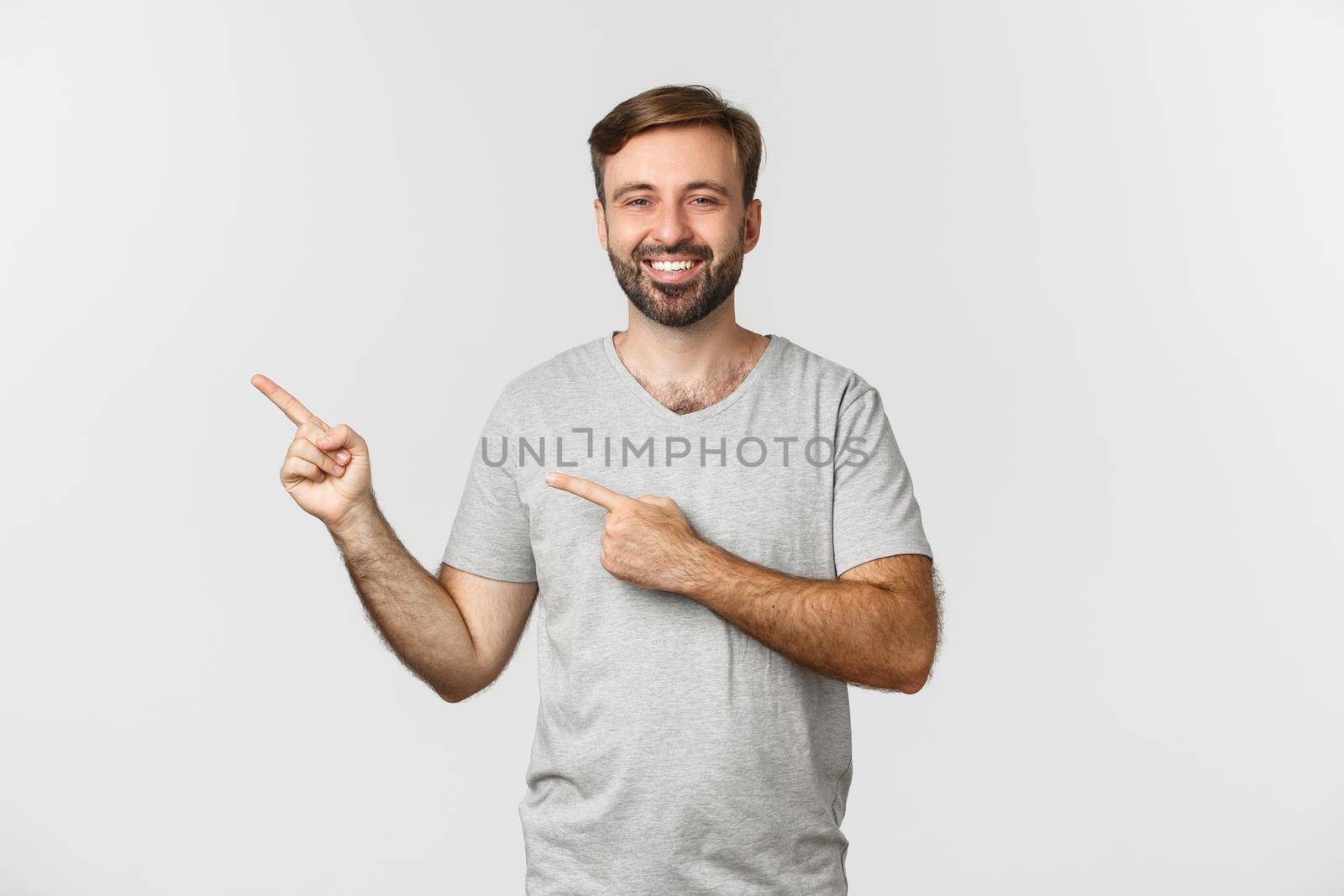 Image of happy guy in casual clothes smiling, pointing fingers left at your logo or promo, standing over white background.