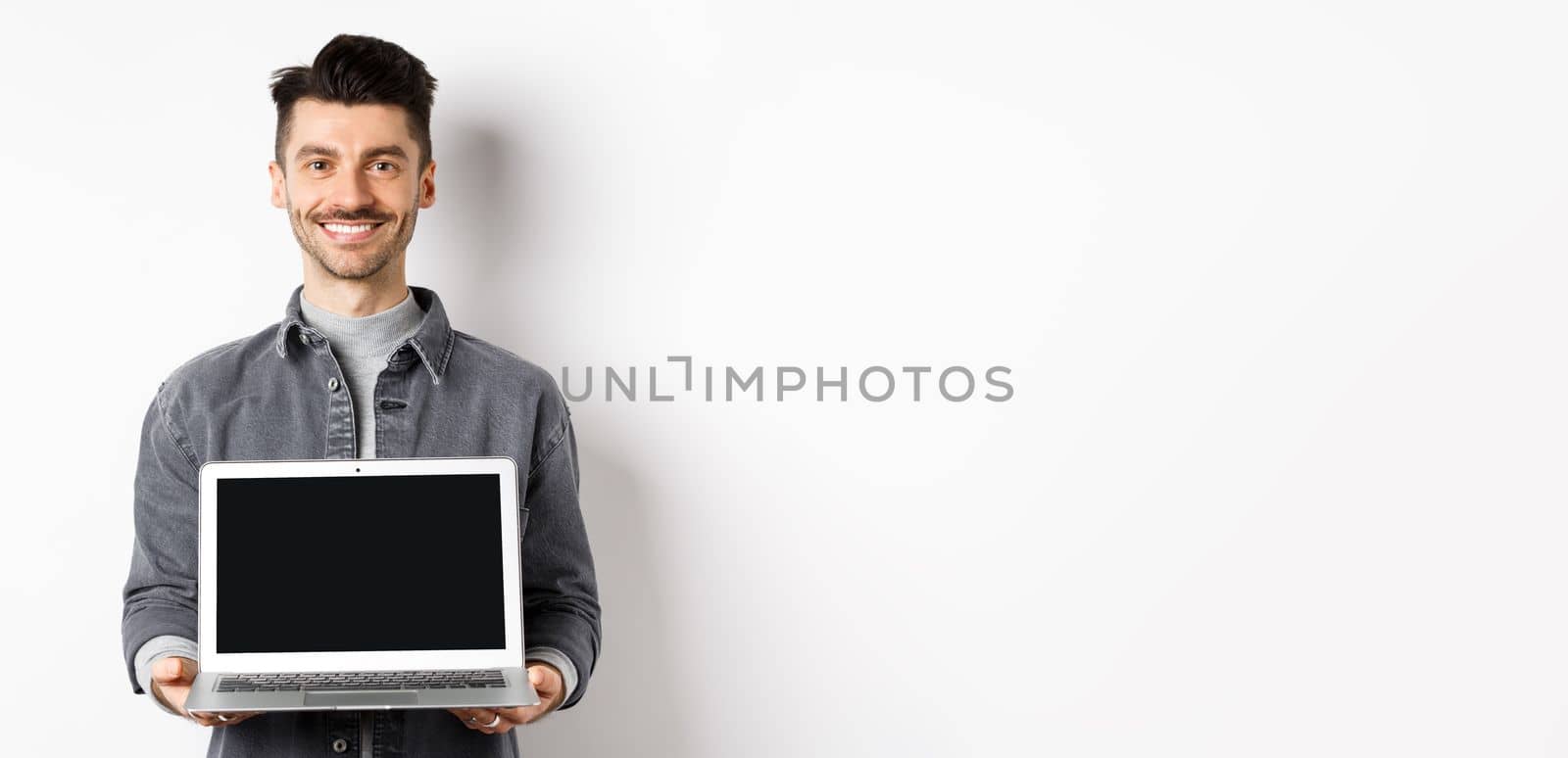 Smiling young man showing empty laptop screen, showing online deal, standing on white background by Benzoix