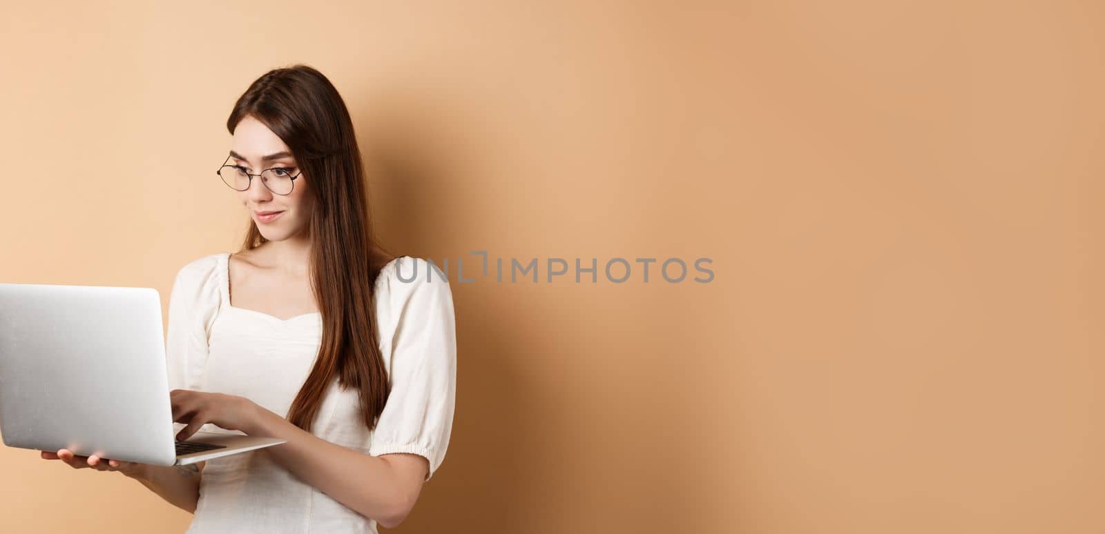Young woman in glasses working on laptop, reading computer screen and smiling, standing on beige background.