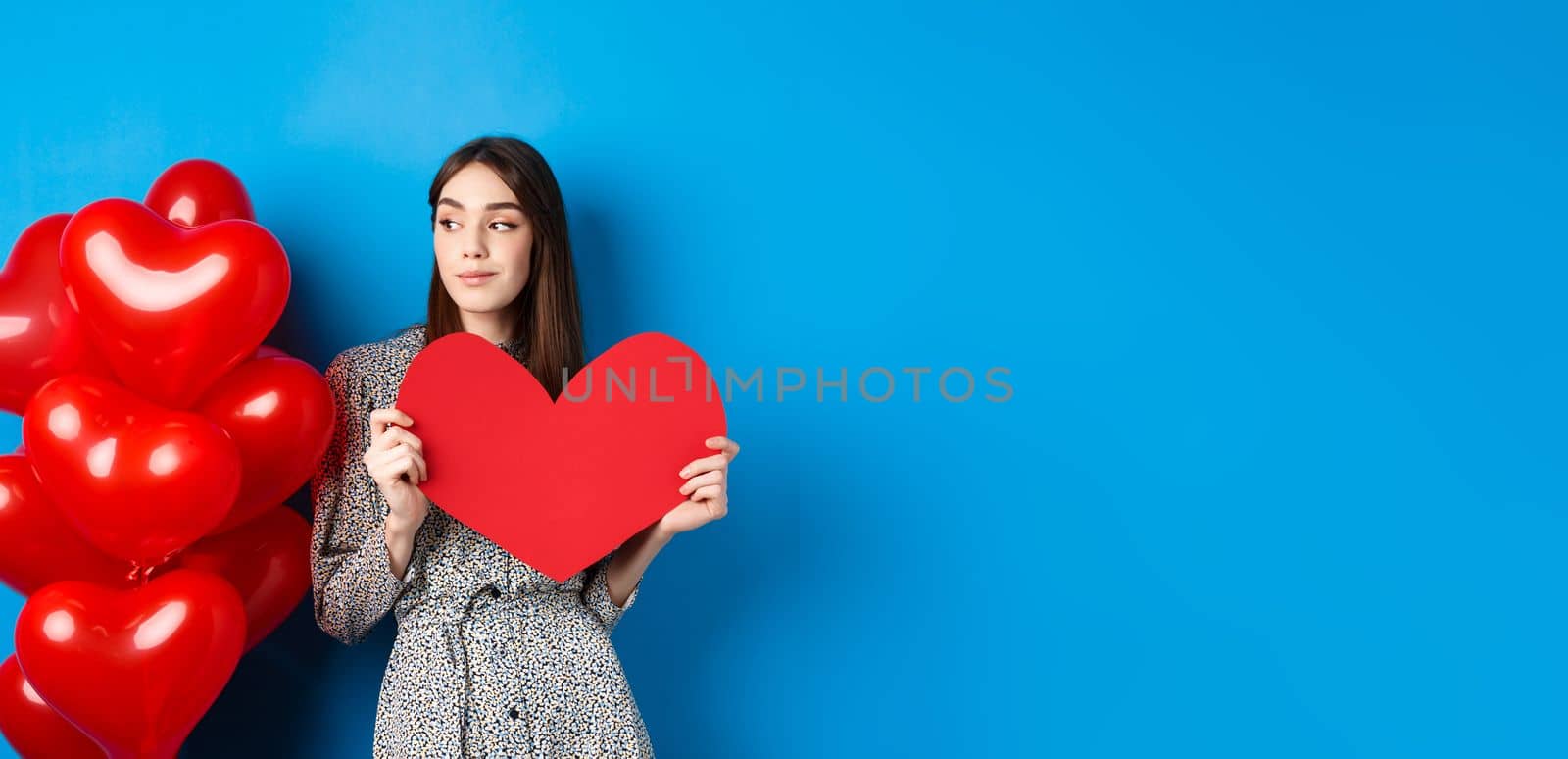 Valentines day. Romantic girl in dress standing near balloons and holding big red heart cutout, dream of something, standing on blue background by Benzoix