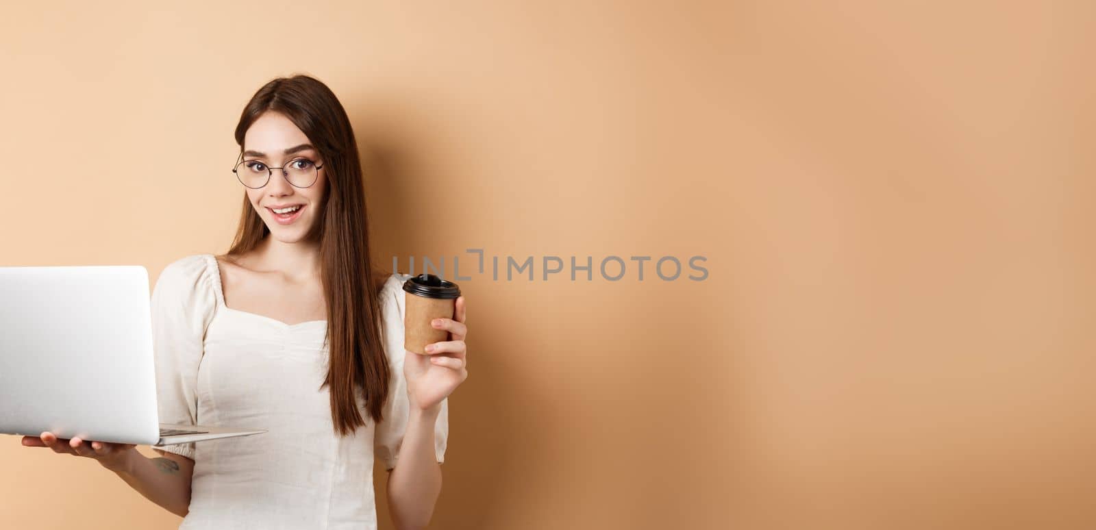 Smiling girl in glasses working on laptop and drinking coffee takeaway. Freelancer using computer at work, standing on beige background by Benzoix