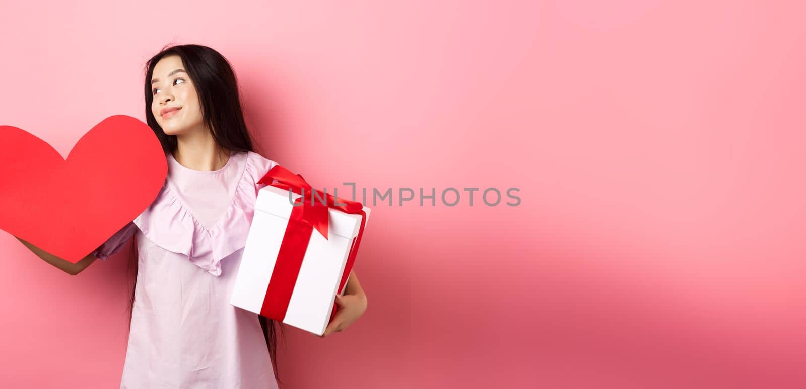 Valentines day concept. Romantic girl holding lover gifts, big red heart card and box with present, looking aside with dreamy look, falling in love, smiling tenderly, pink background.