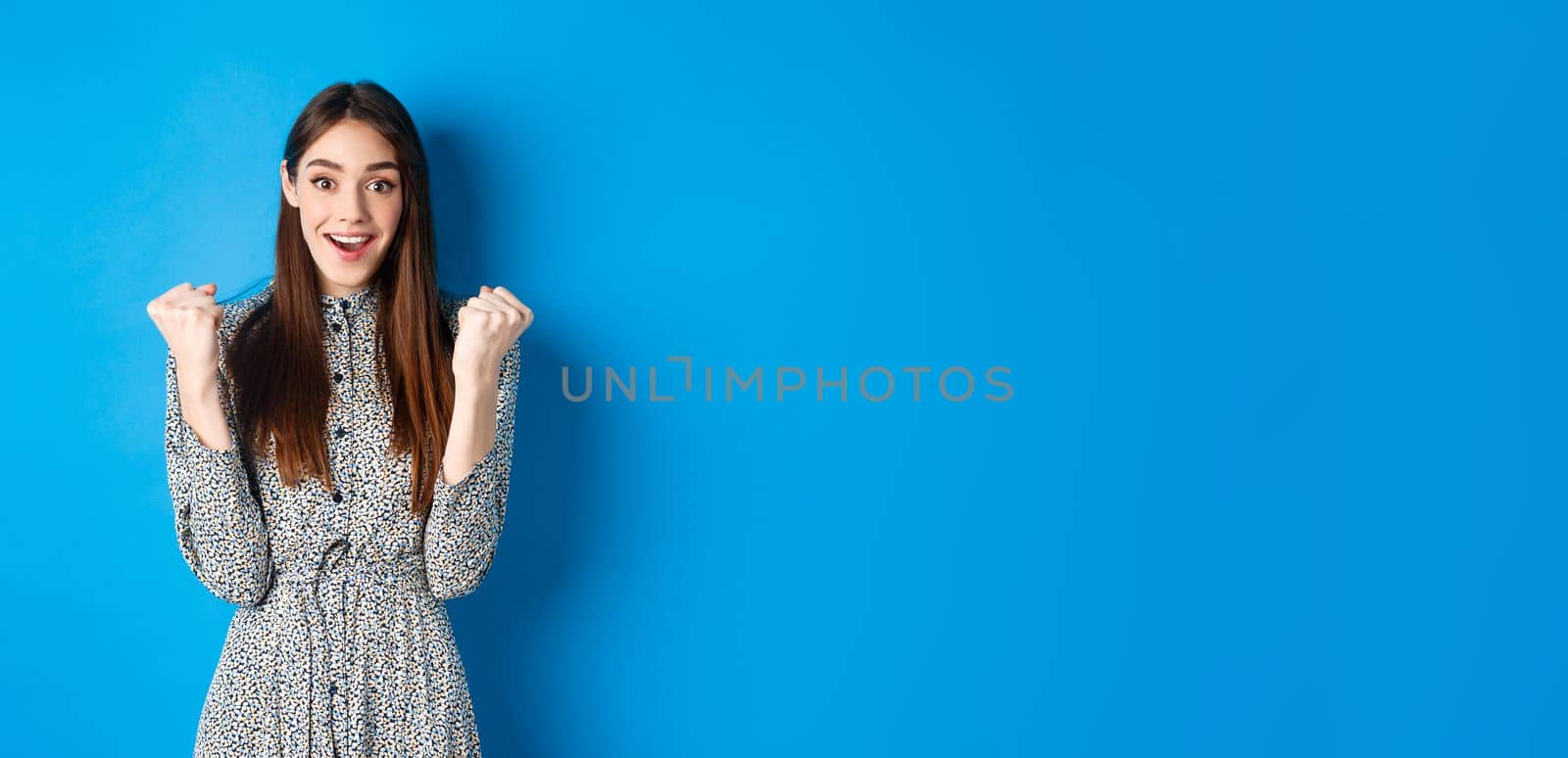 Excited woman looking relieved and hopeful, making fist pump and smiling, winning prize, celebrating achievement or victory, standing on blue background by Benzoix