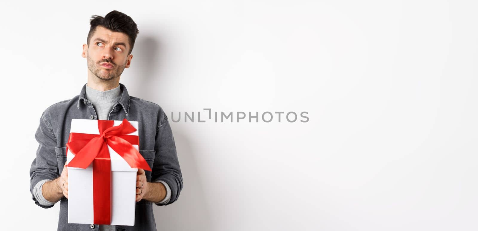 Pensive boyfriend looking aside and holding gift box, waiting for lover, making surprise present on valentines day, planning date with girlfriend, white background by Benzoix