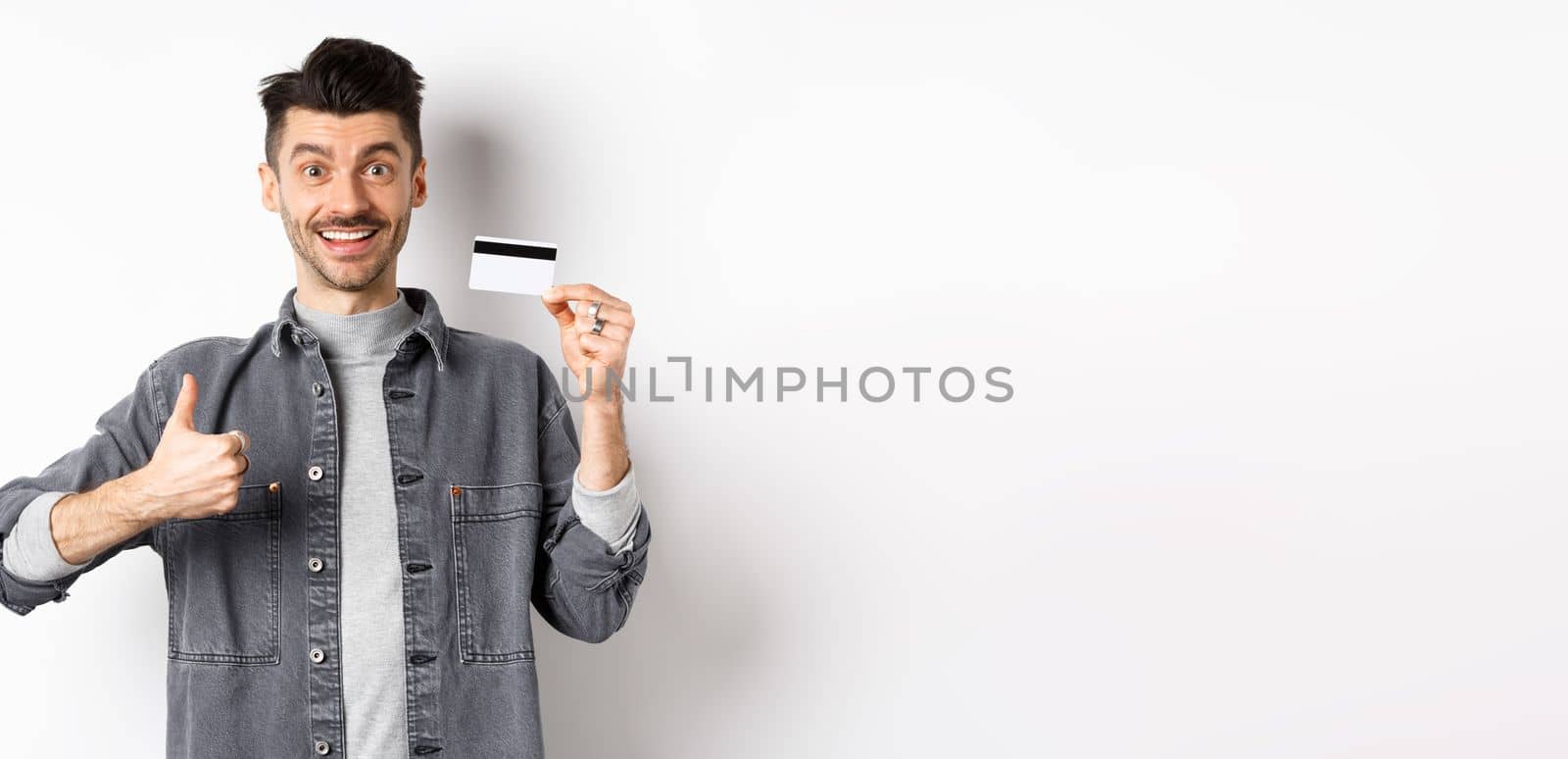 Very good. Smiling guy with plastic credit card showing thumbs up, smiling satisfied, recommend bank, standing on white background.