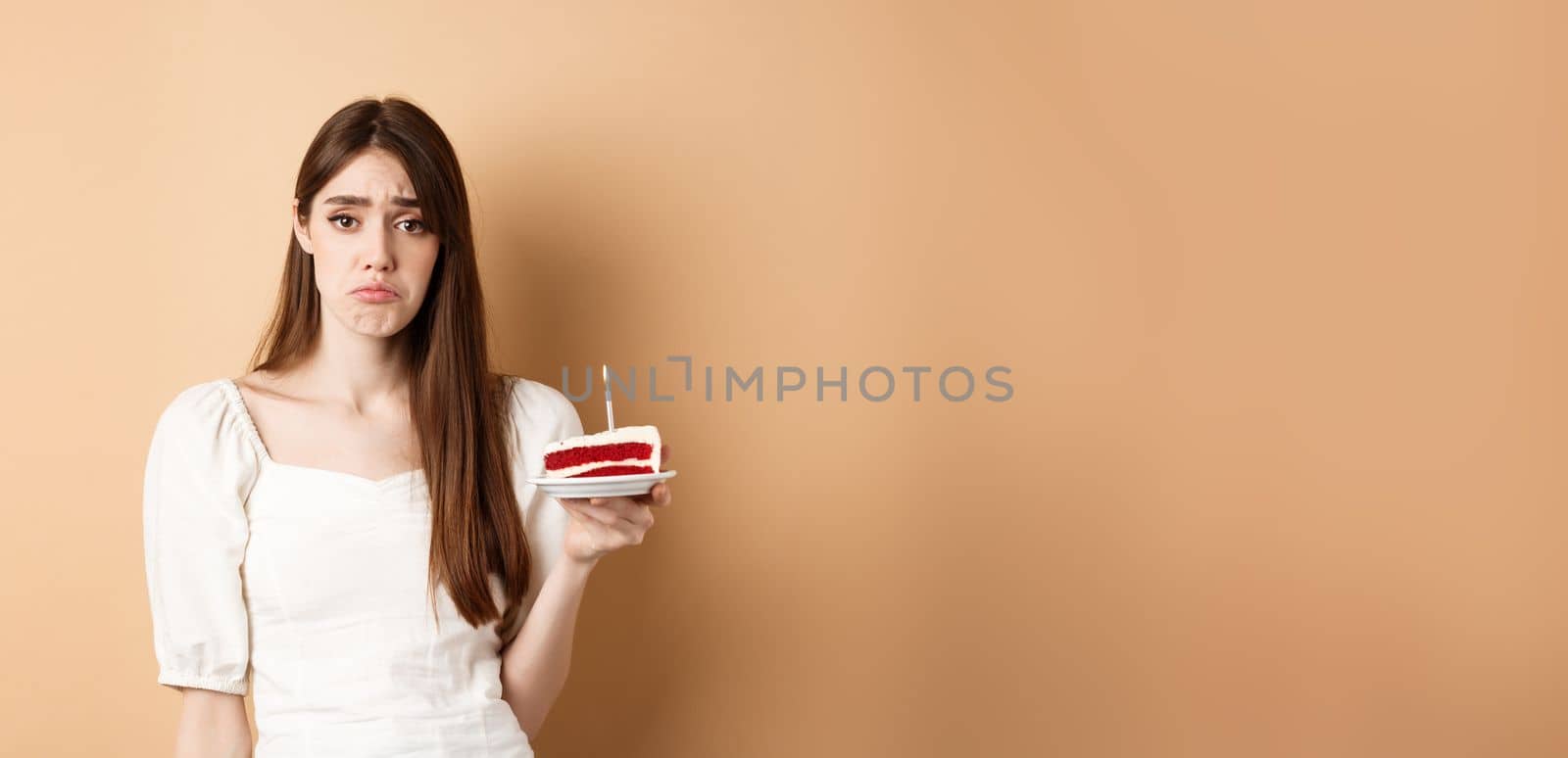 Sad birthday girl holding cake with candle and look upset, feeling lonely on her bday, standing alone on beige background by Benzoix