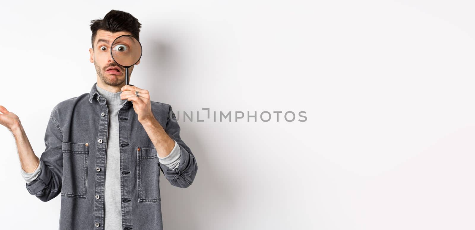 Confused man look through magnifying glass and shrugging clueless, standing on white background.