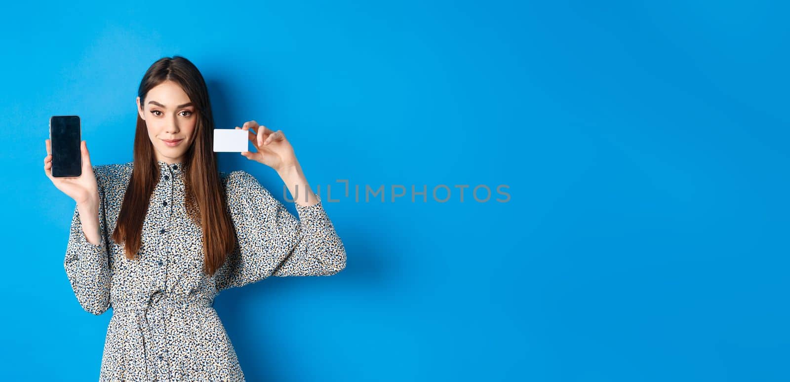 Online shopping. Confident beauty woman showing plastic credit card and empty smartphone screen, showing stop, standing on blue background by Benzoix
