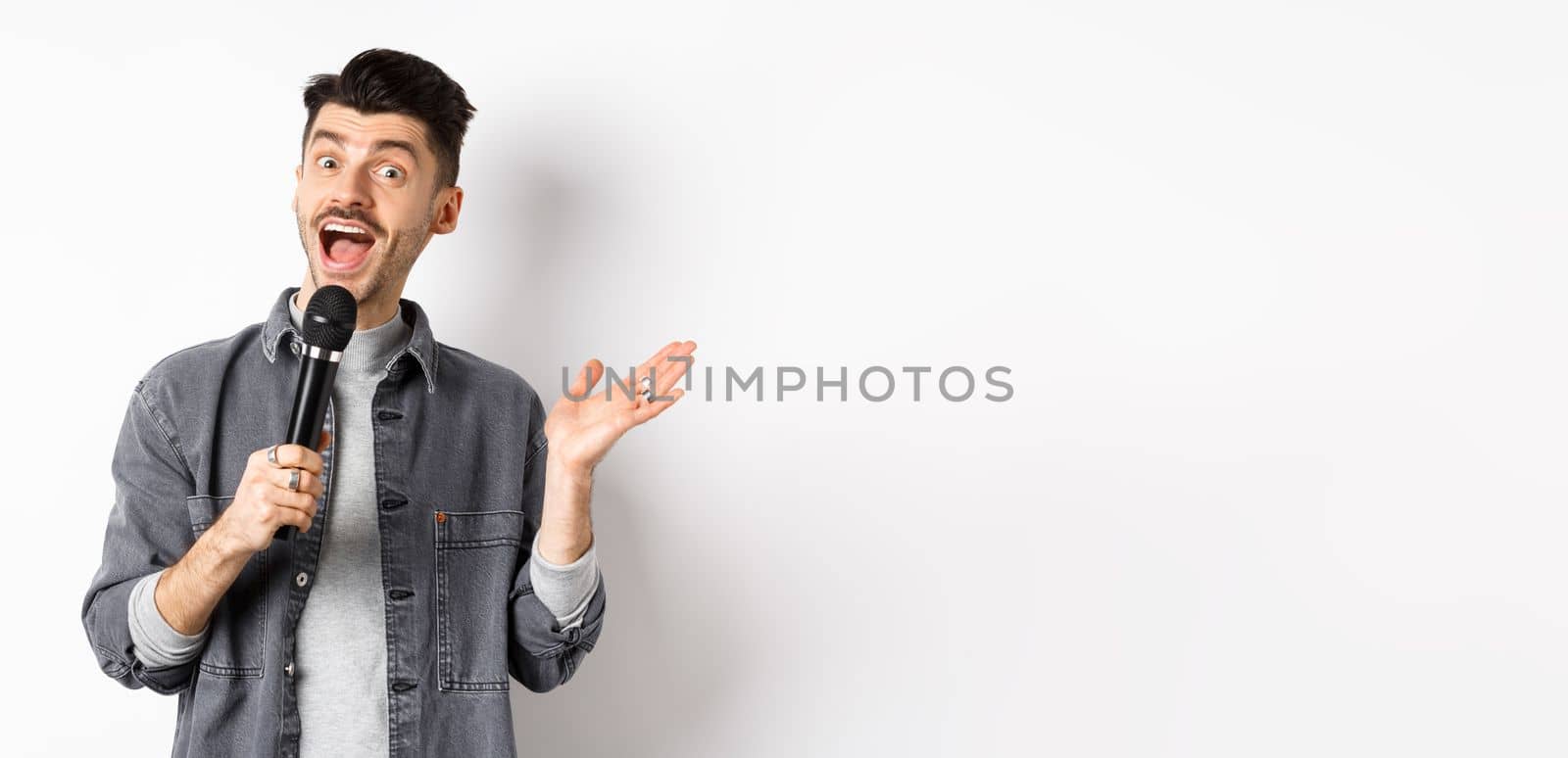 Excited stylish guy giving speech, talking in microphone, perform or sing karaoke, standing on white background.