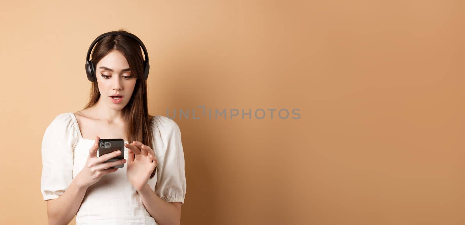 Excited girl listen music in headphones and looking at smartphone amazed, reading awesome news, found cool playlist, standing on beige background.