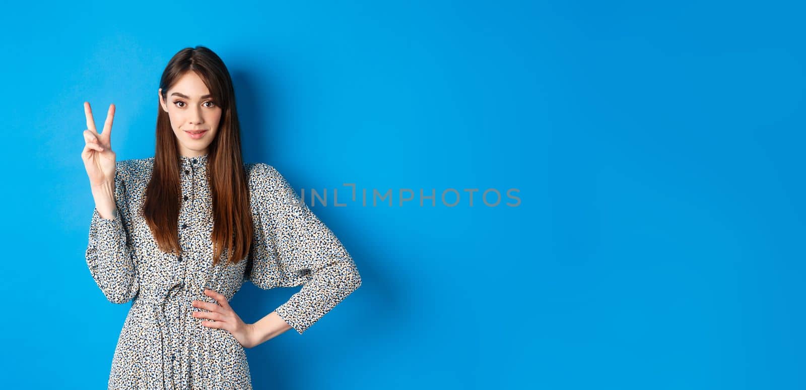 Young beautiful girl in dress with natural long hair, showing number two with fingers and smiling, standing against blue background.
