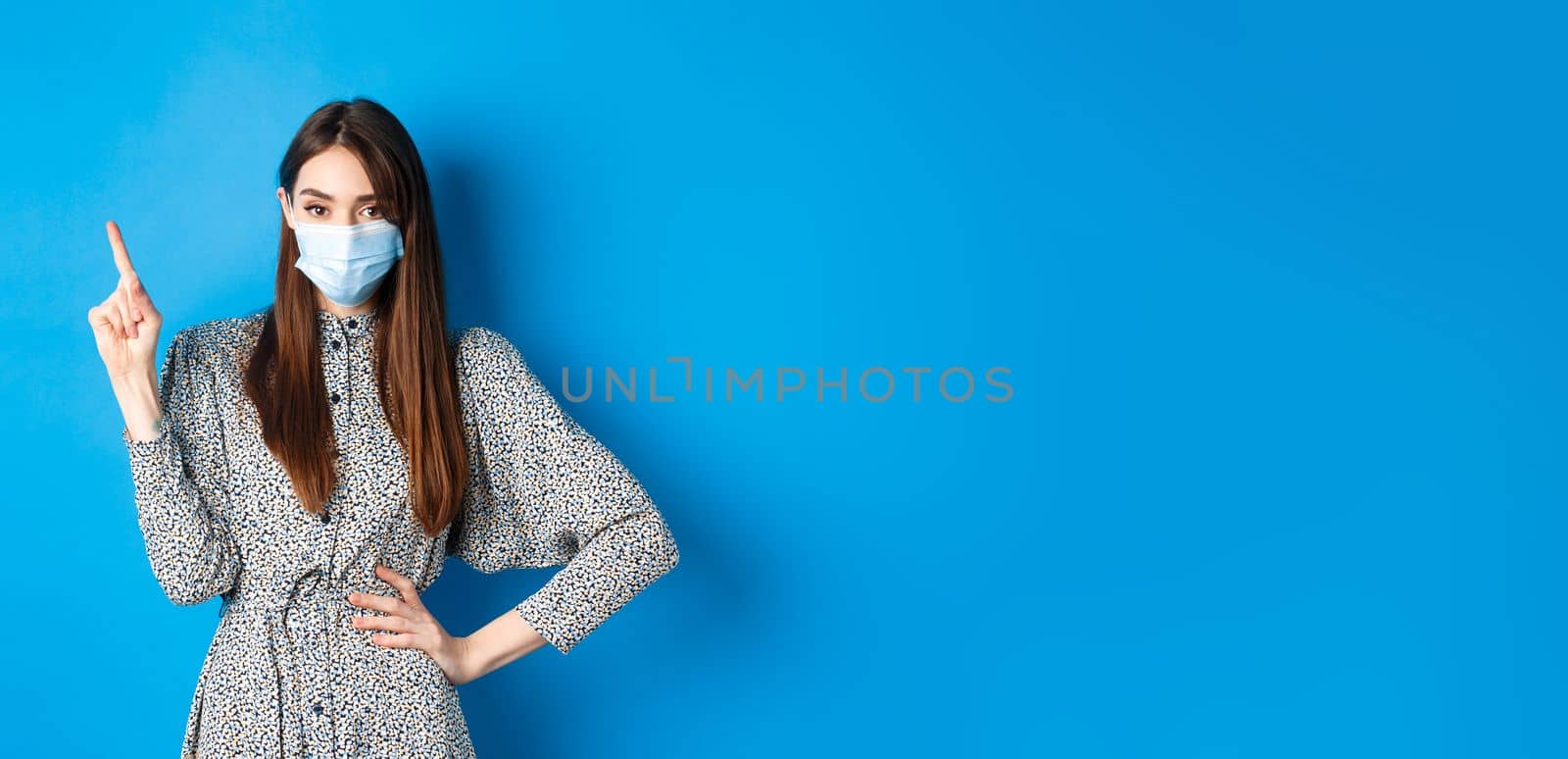 Healthy people and covid-19 pandemic concept. Serious woman in medical mask pointing at upper left corner, standing on blue background by Benzoix