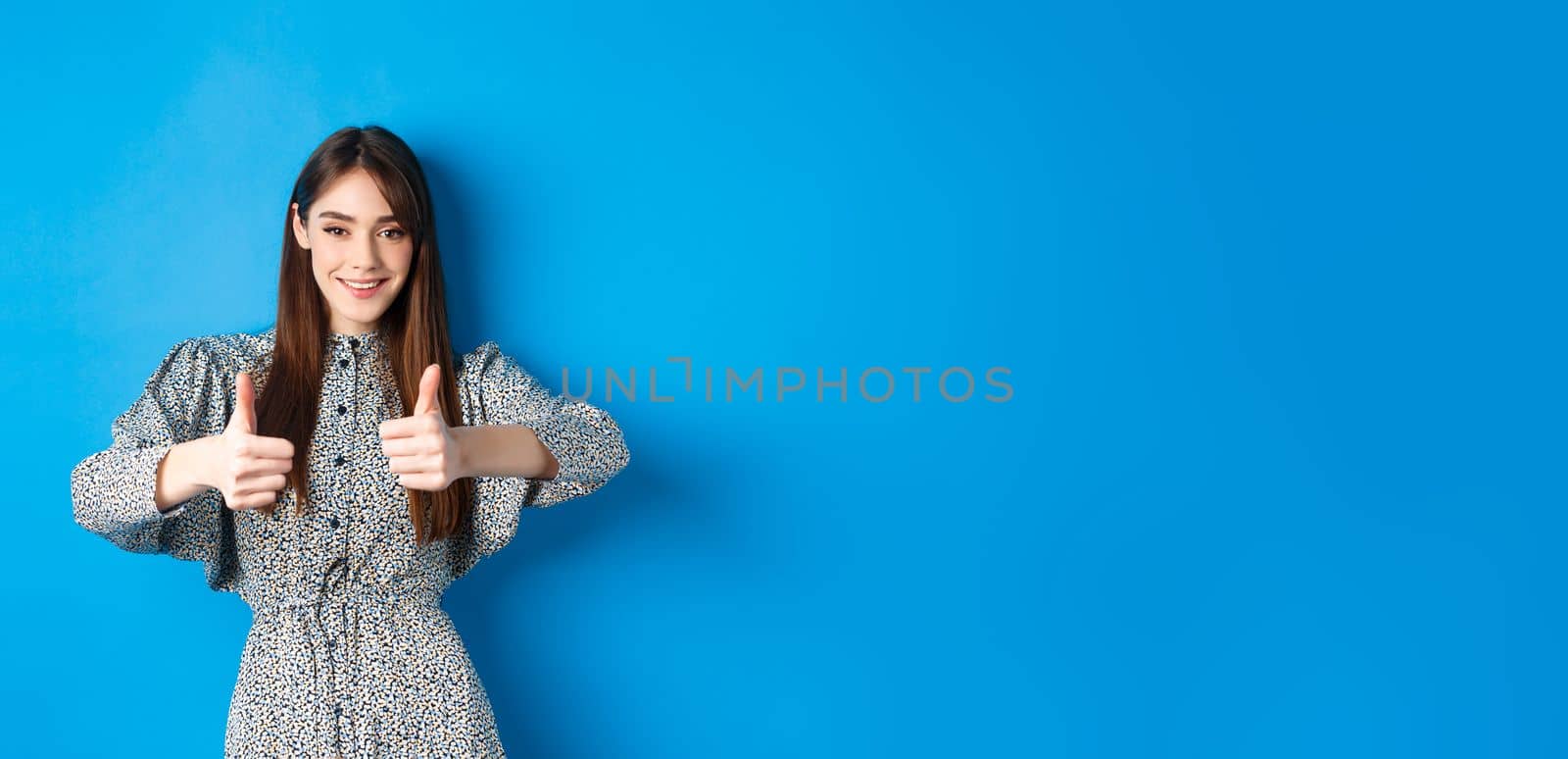 Nice work, very good. Smiling young woman showing thumbs up to support your choice, looking pleased and approve something, standing in dress against blue background by Benzoix