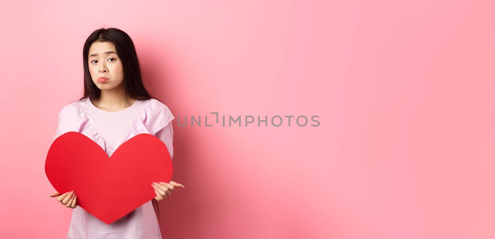 Valentines concept. Single teenage asian girl wants to fall in love, looking sad and lonely at camera, sulking distressed on lovers day, holding big red heart cutout, pink background.