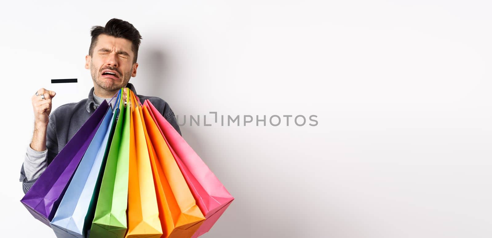 Sad crying man wasted all money on shopping, holding bags and showing empty credit card, standing against white background.