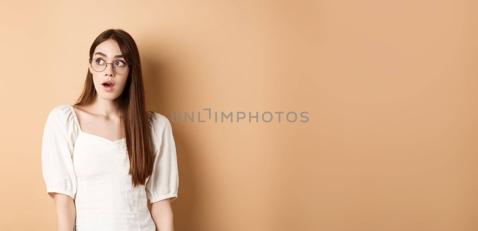 Excited young woman in glasses looking aside with opened mouth, gasping fascinated, standing on beige background by Benzoix
