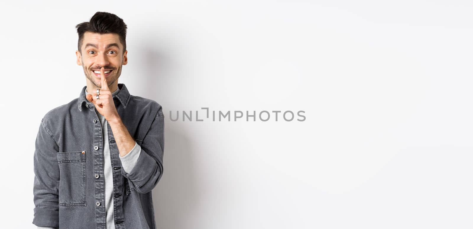 Happy guy shushing at camera, smiling with finger on lips, asking to keep quiet, telling big secret or making surprise, standing on white background.