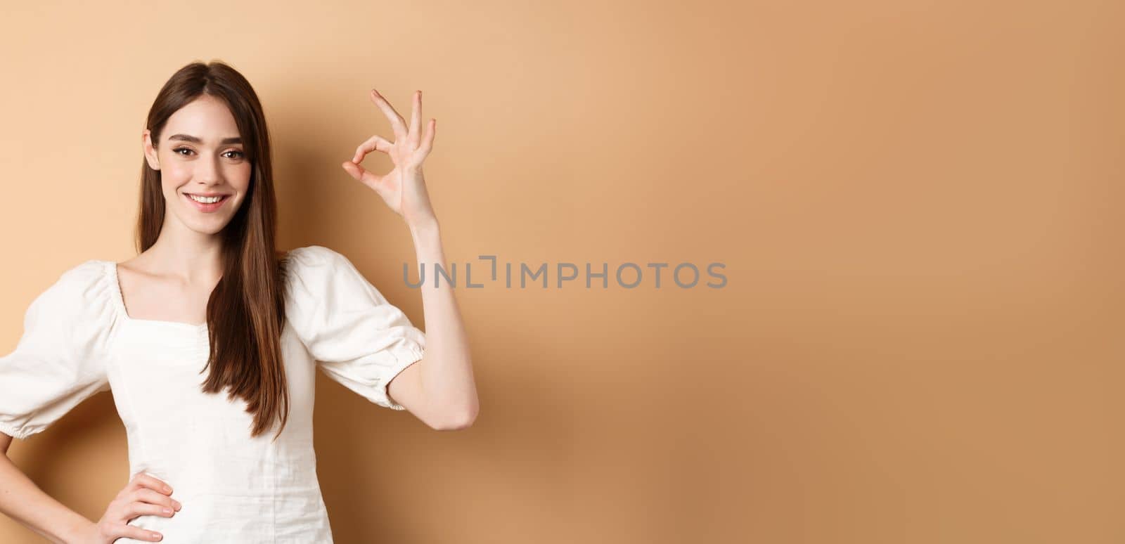 Very good. Smiling cute woman in white blouse showing okay sign in approval, like and praise excellent choice, standing satisfied on beige background.