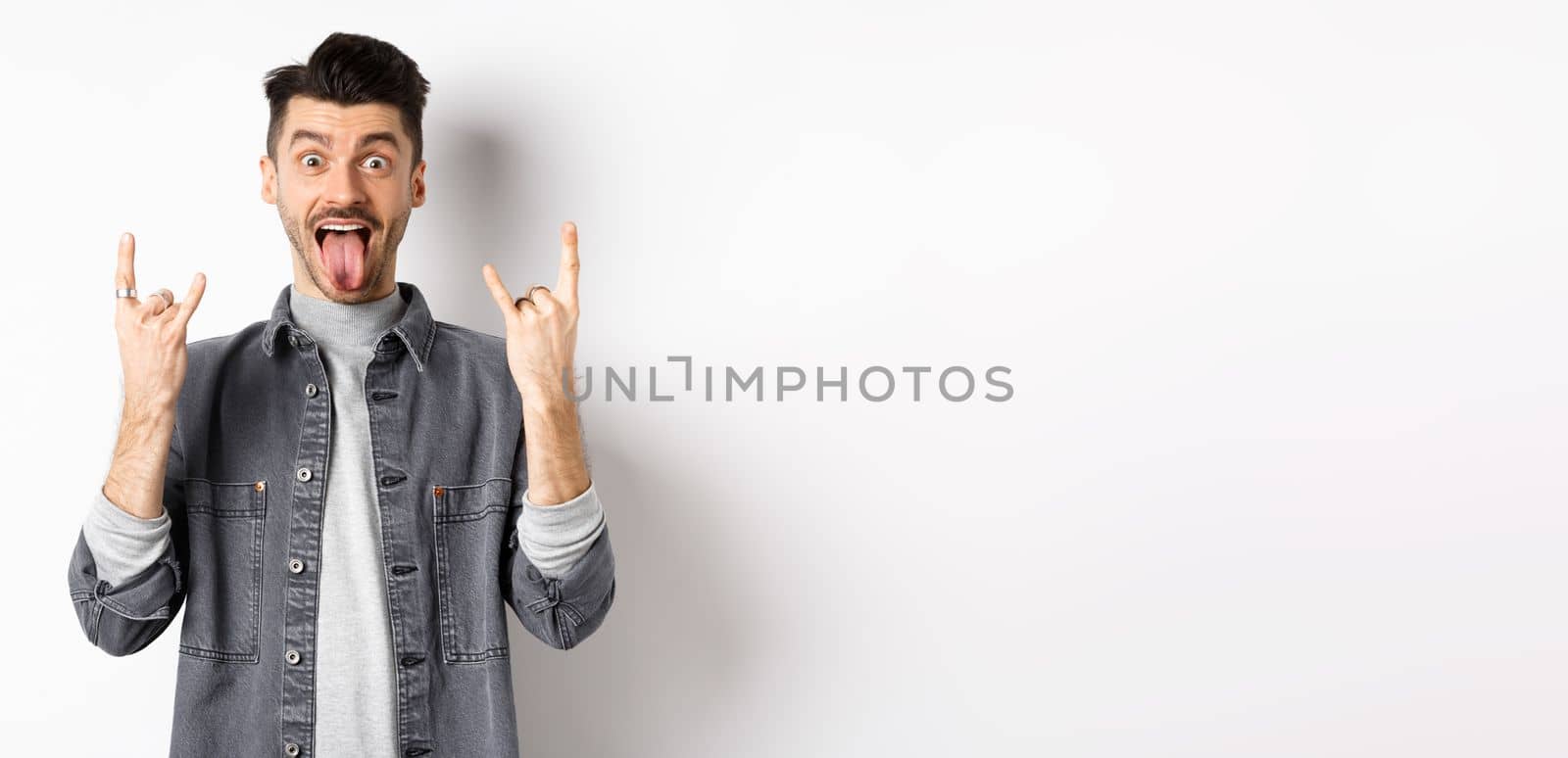 Excited funny guy showing tongue and heavy metal horns sign, enjoying party or event, having fun, standing on white background.