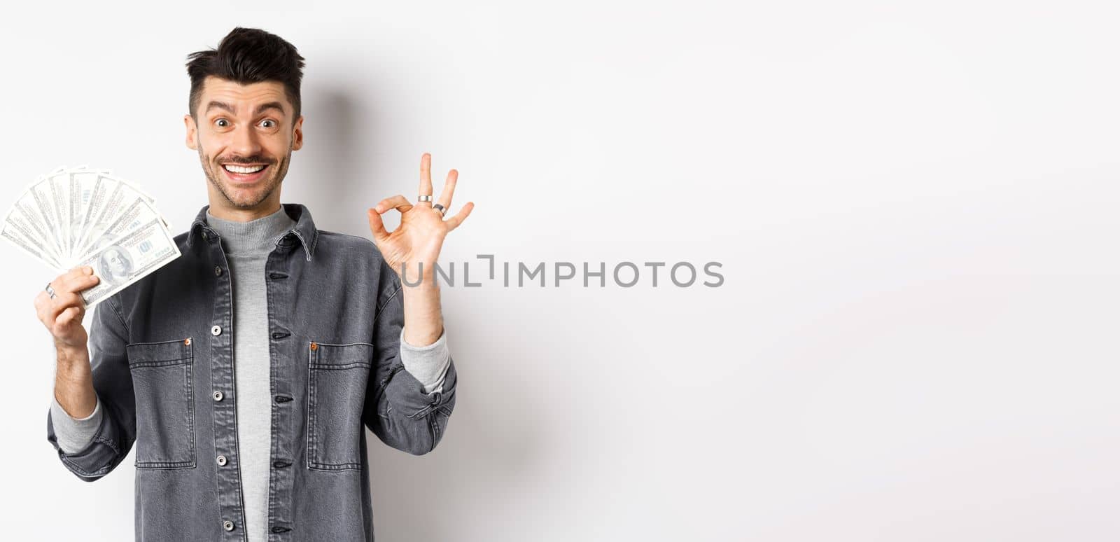 Image of excited man holding dollar bills and show okay sign with happy face, making money, standing on white background.