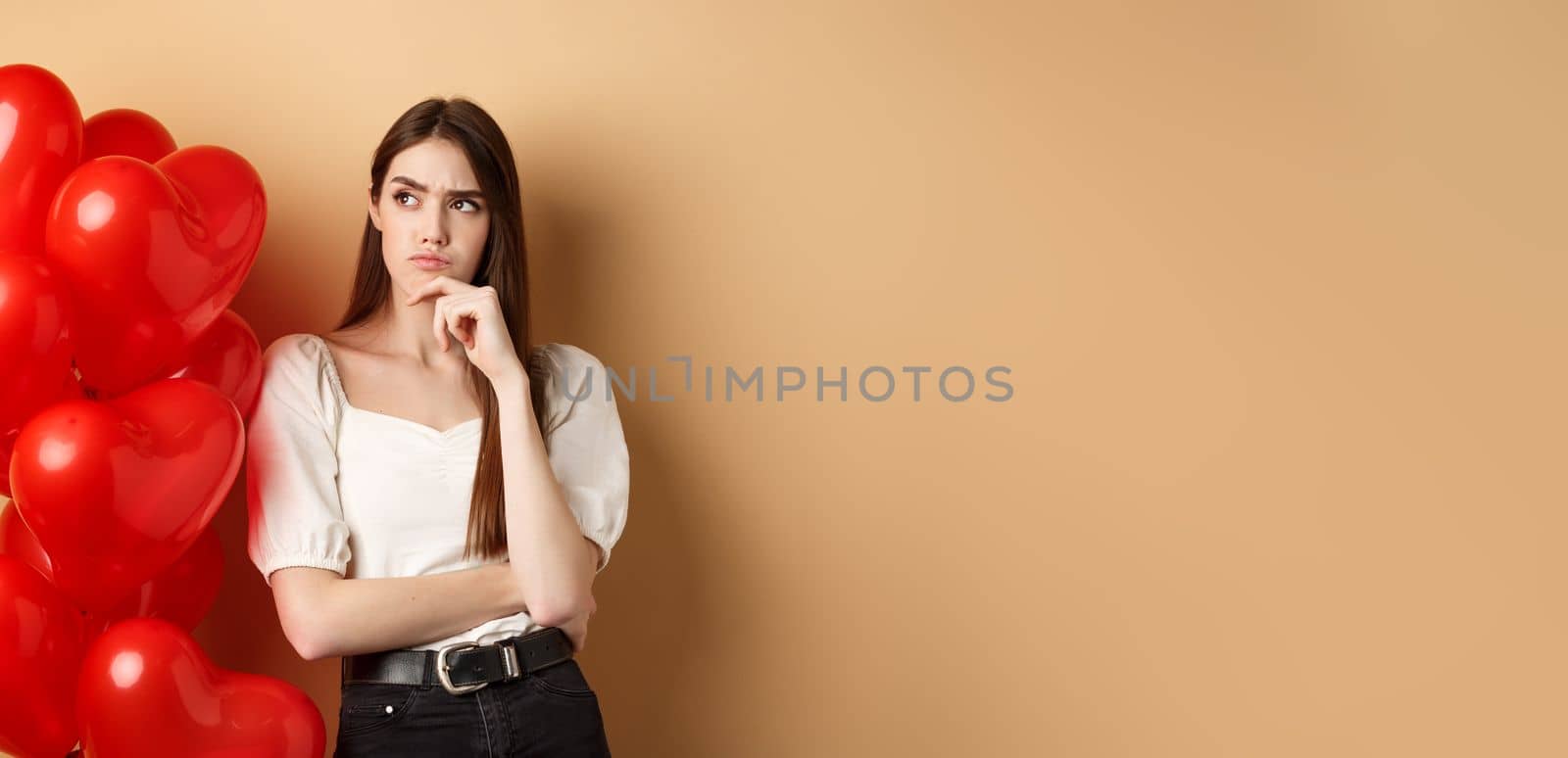Valentines day and love concept. Thoughtful young woman standing near hearts balloons and looking aside with doubtful face, having assumptions, beige background.