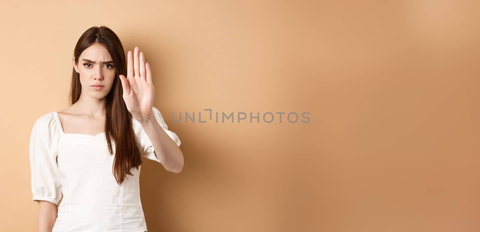 Nope stop right there. Serious and confident woman stretch out hand to forbid something, frowning and saying no, disagree and reject bad offer, standing on beige background.