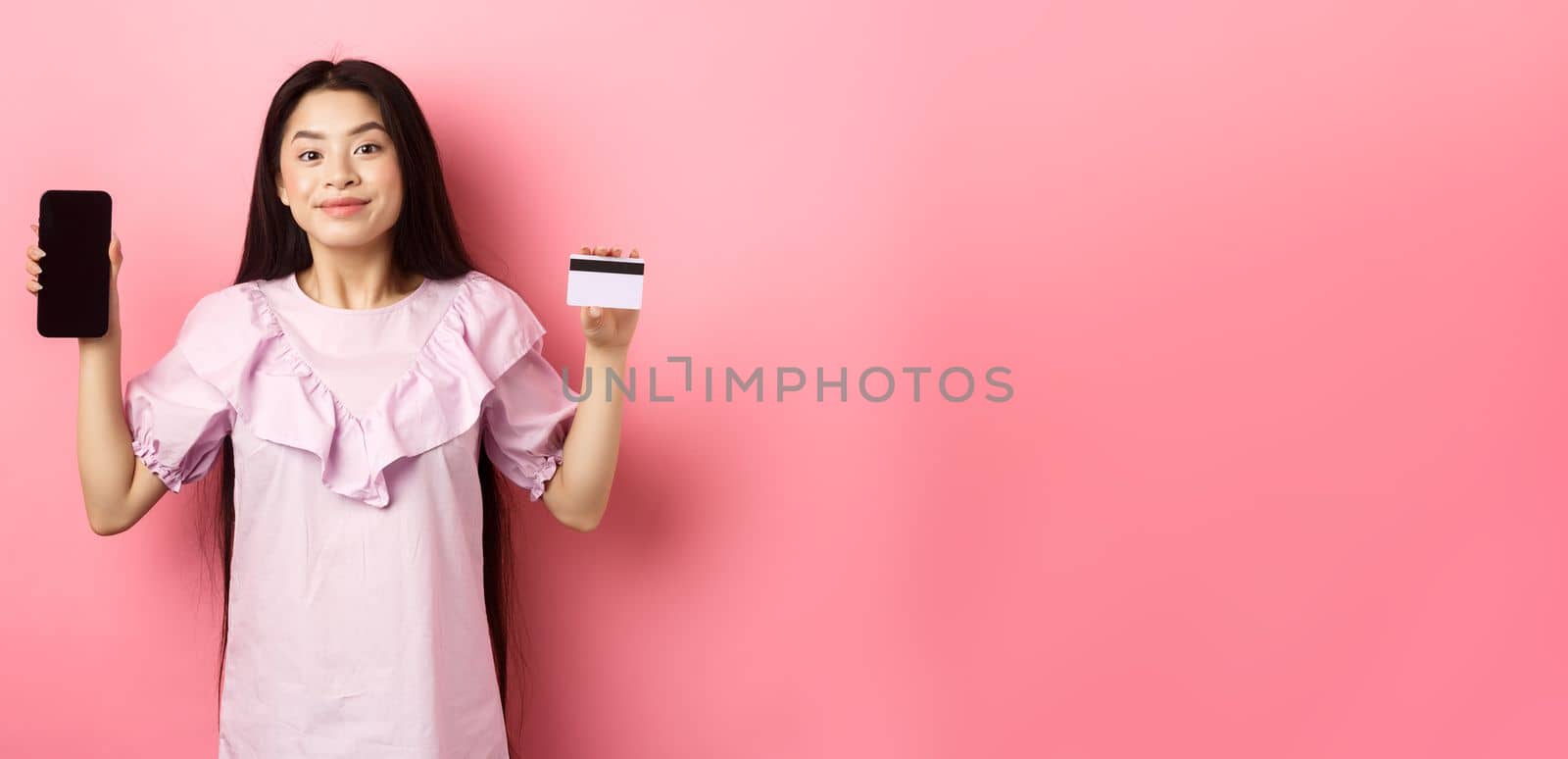 Cheerful asian girl showing empty smartphone screen and credit card, shopping online, standing on pink background by Benzoix