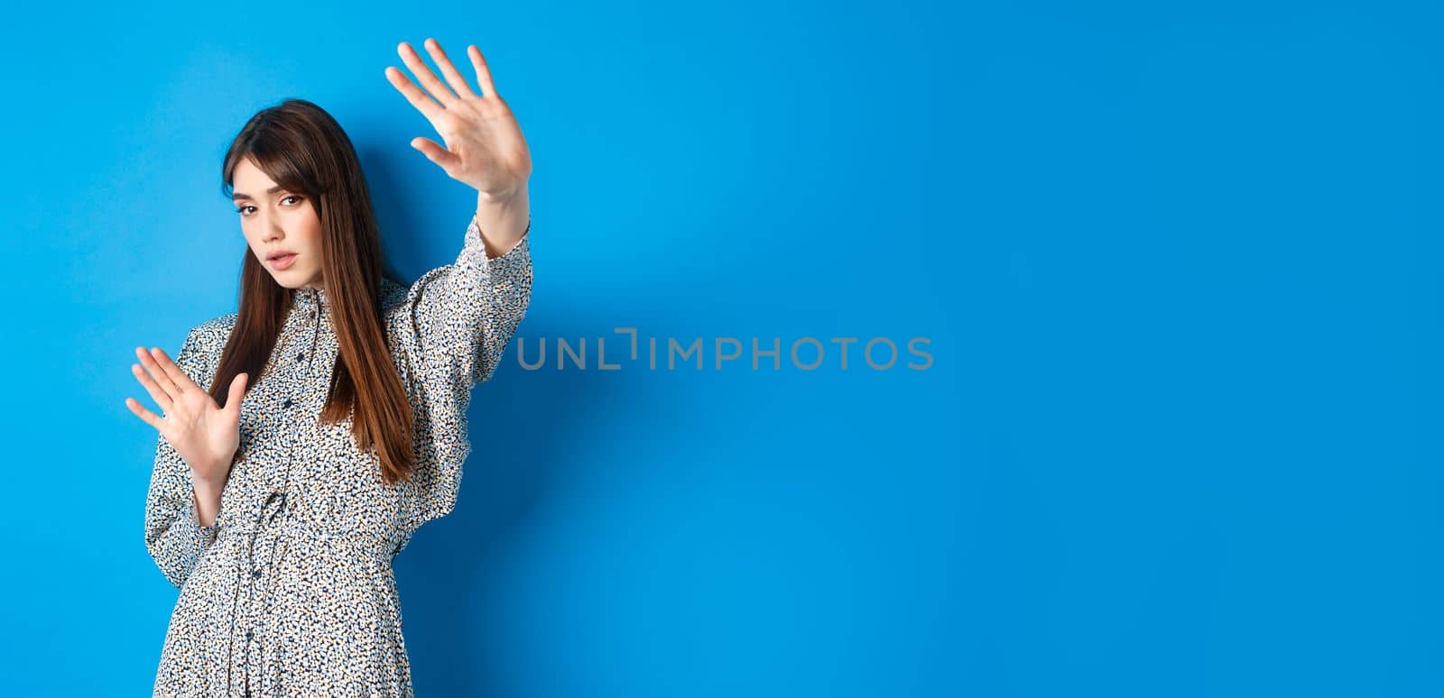 Image of young woman raising hands up defensive, covering herself from camera fleshlights, standing on blue background. Copy space