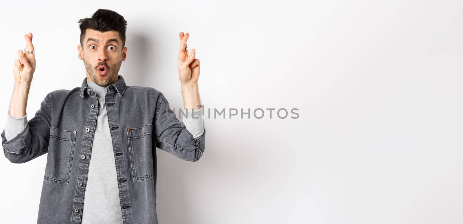 Excited young man cross fingers for good luck and gasping, holding breath as waiting important news, begging or praying, standing on white background.