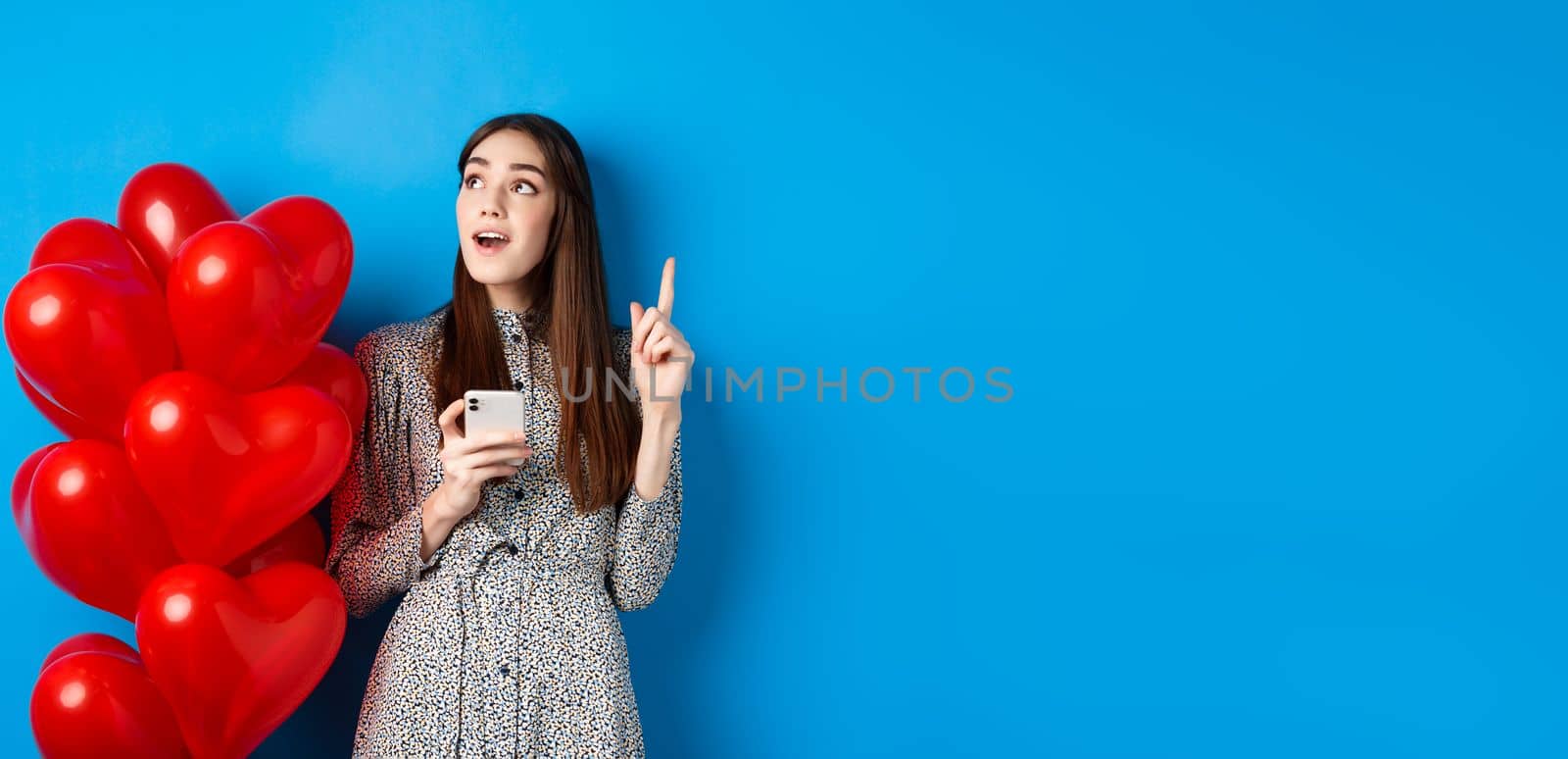 Valentines day. Image of romantic girl pitching an idea after using mobile phone, raising finger up and looking at empty space, standing near red heart balloons, blue background by Benzoix