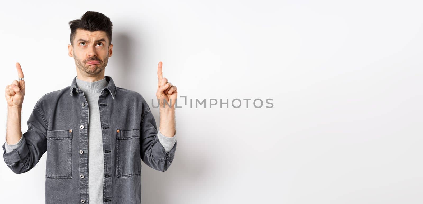 Sad frowning guy pointing and looking up, standing hesitant on white background.