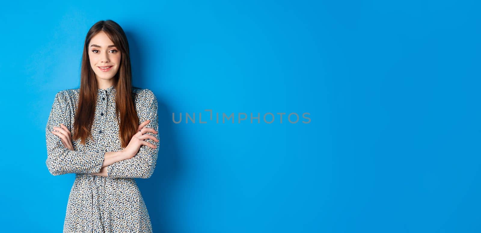 Elegant caucasian girl in dress cross arms on chest, standing on blue background.