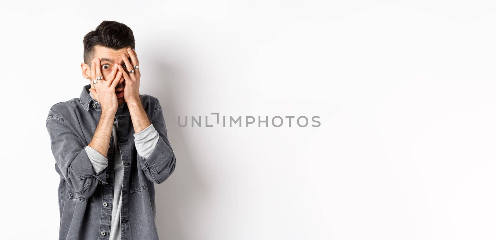 Scared young man peeking through fingers with nervous face, staring at something scary, standing on white background.