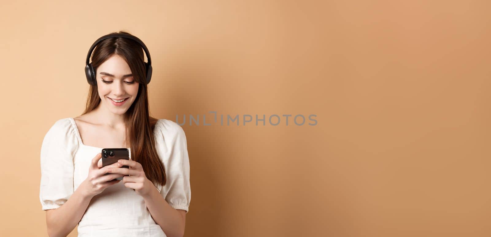 Smiling woman texting message on phone and listening music in wireless headphones, watching video on smartphone, beige background.