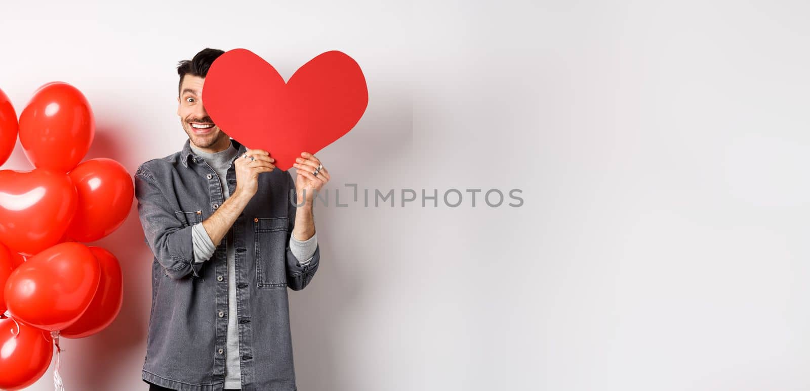 Romantic smiling man cover face with Valentine heart card and looking happy at camera, celebrating lovers day with partner, standing on white background.