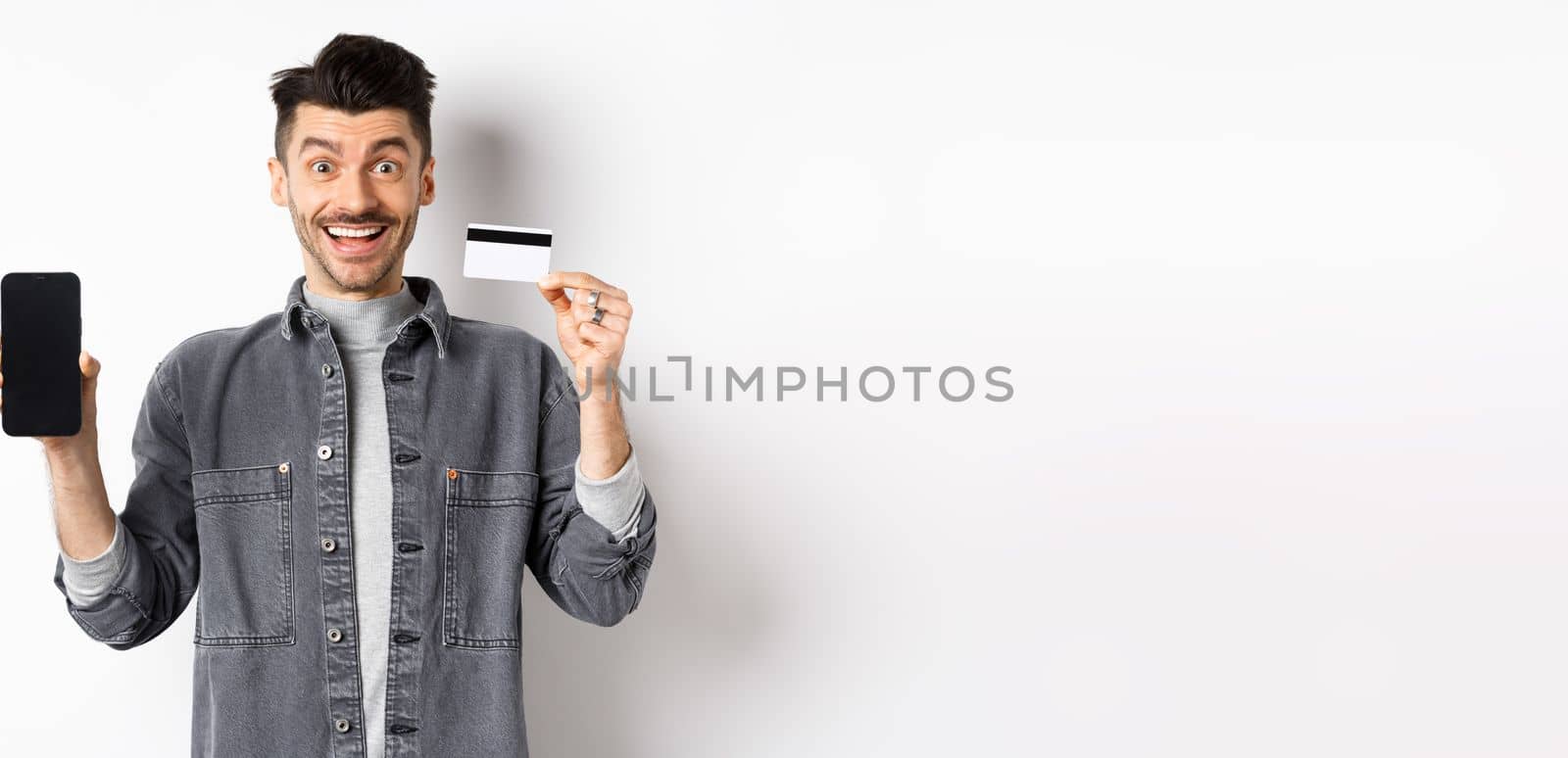 Excited handsome man showing plastic credit card and mobile phone screen, standing on white background by Benzoix