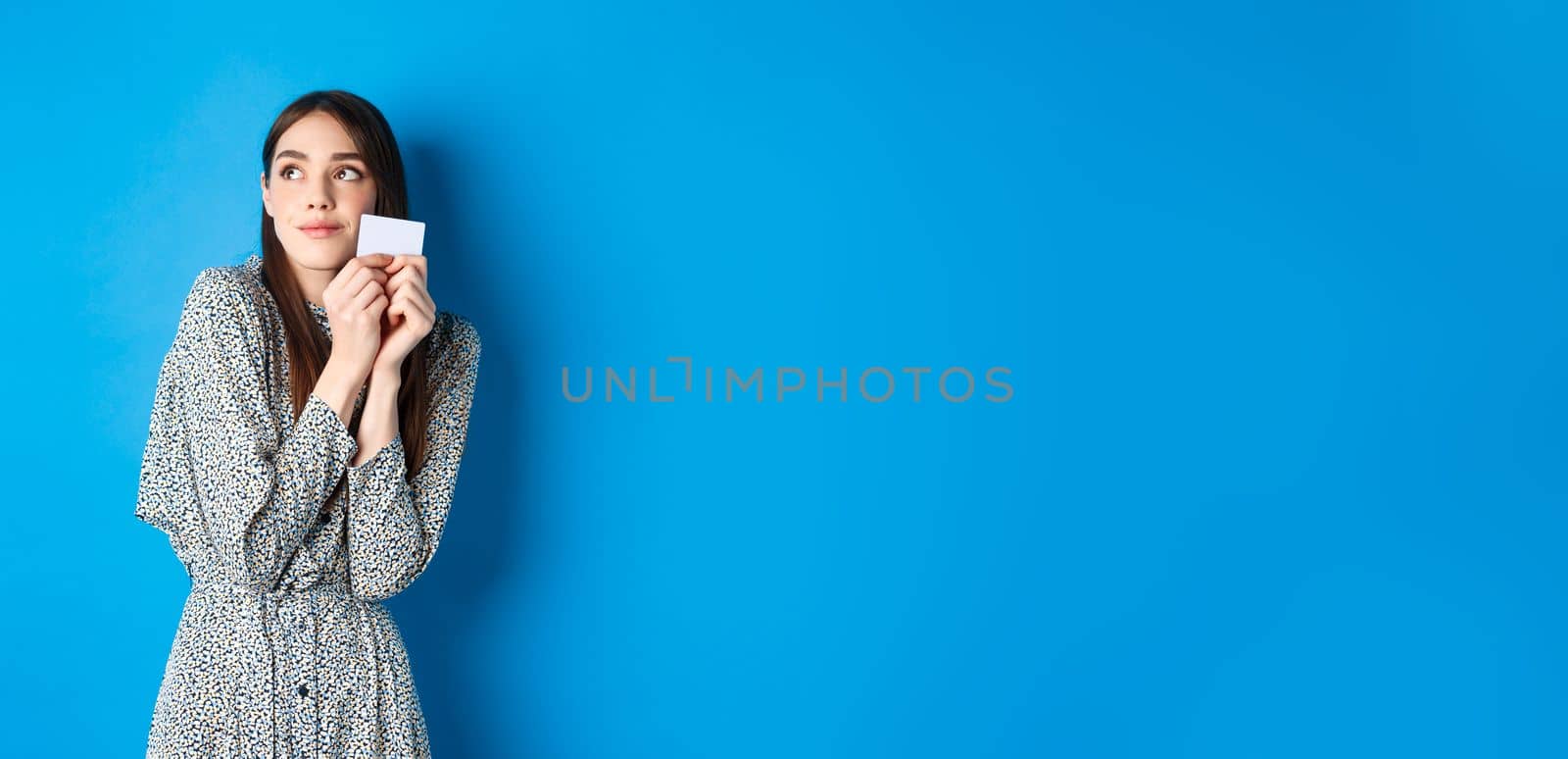 Romantic cute girl in dress dreaming of shopping, holding credit card and looking at logo with tender smile, standing on blue background by Benzoix