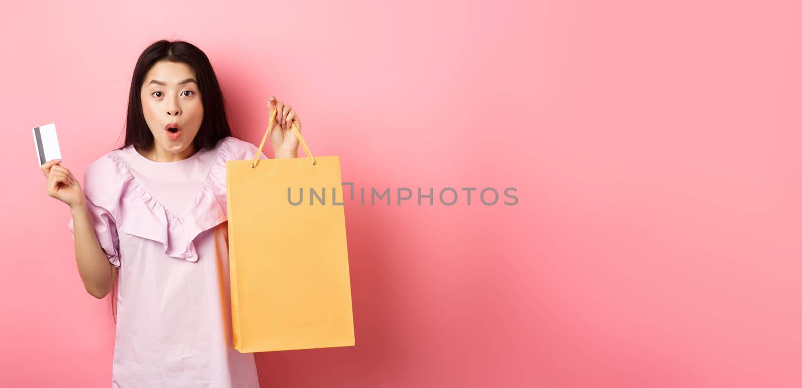 Shopping. Excited asian woman gasping amazed, holding bag from shop and plastic credit card, standing on pink background.